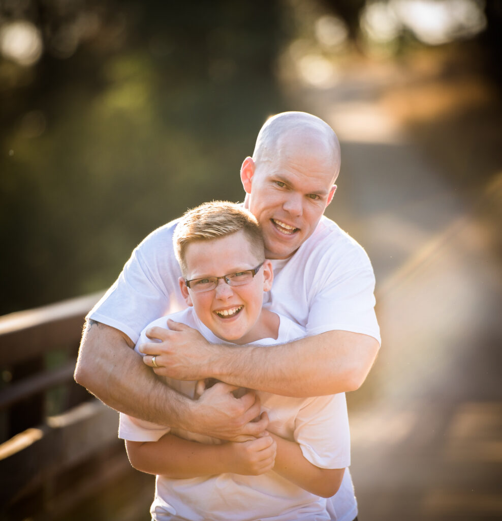 Portrait of a father embracing his son on a bridge at sunset, while both of them are smiling and happy.