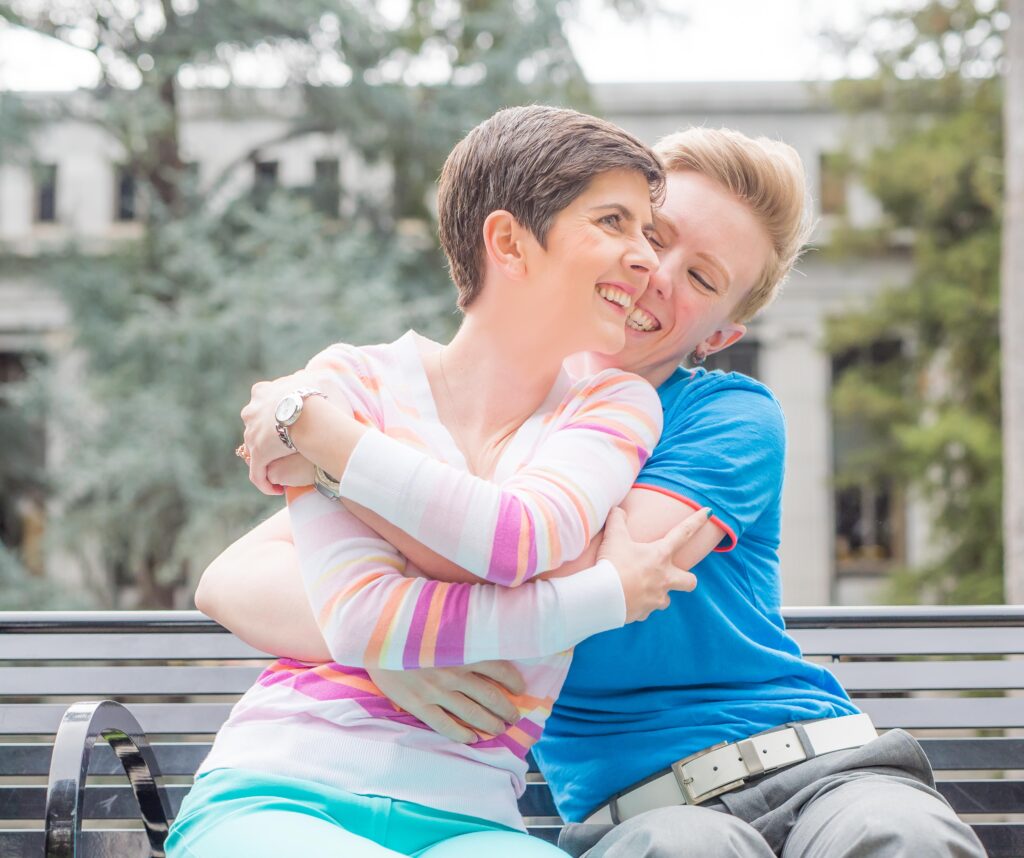 Portrait of a female couple embracing on a bench in a park looking at each other and off into the distance.