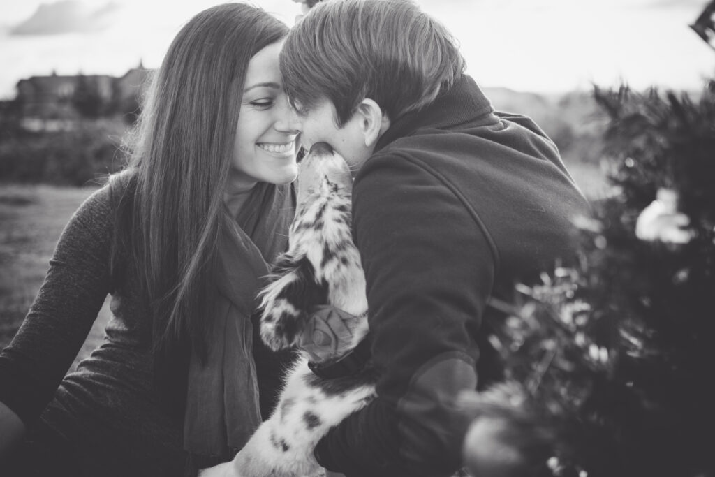 Portrait of a female couple in black and white with their dog. One woman is looking toward the camera and smiling, while the other is looking away with her dog kissing her.