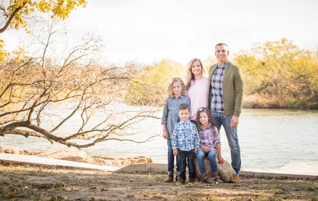 Portrait of a family of five posing in front of a river near a tree during the daylight.