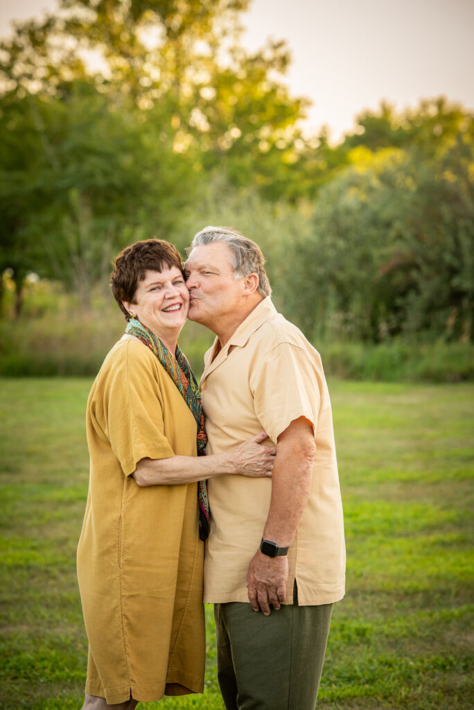 Portrait of an older couple in a tree filled area embracing each other with the man kissing the woman on the cheek, while both are happy and smiling.