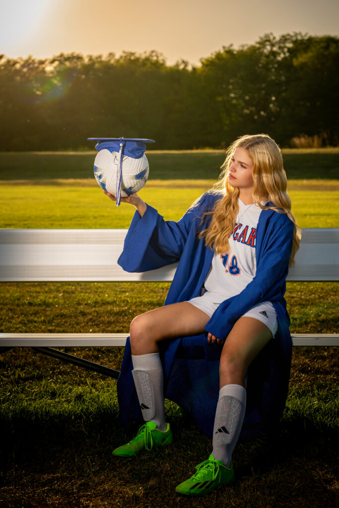 High school senior in soccer uniform and cap and gown holding a soccer ball during sunset