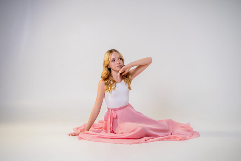 A young dancer in a pink skirt and white leotard sitting on a studio floor